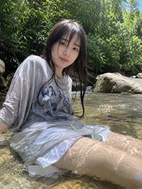 A person in a loose gray outfit sits in clear shallow water surrounded by rocks and leafy trees.