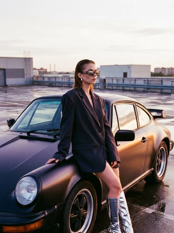 Woman in a dark structured jacket beside a vintage car at sunset